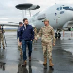 Ο αντιπρόεδρος των ΗΠΑ με σκούρο μπουφάν μπροστά σε στρατιωτικό αεροσκάφος επιτήρησης με ραντάρ στη ράχη