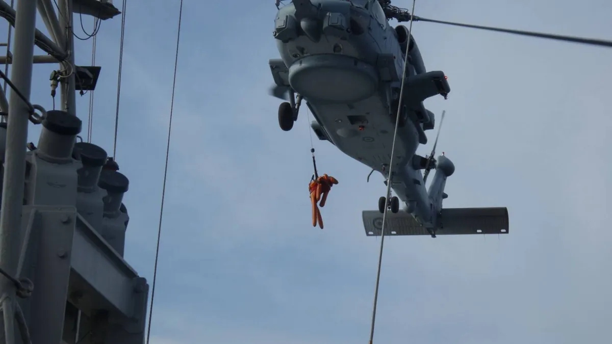 Γκρίζο ναυτικό ελικόπτερο αιωρείται πάνω από το κατάστρωμα κανονιοφόρου ενώ στέλεχος με πορτοκαλί στολή κρέμεται από το συρματόσχοινο