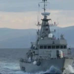 Γκρίζα κανονιοφόρος του Πολεμικού Ναυτικού πλέει κατά μέτωπο με ταχύτητα, με βουνά στο βάθος