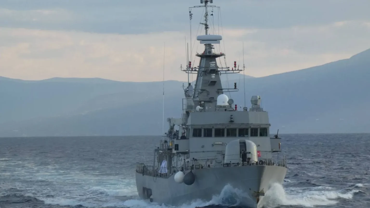 Γκρίζα κανονιοφόρος του Πολεμικού Ναυτικού πλέει κατά μέτωπο με ταχύτητα, με βουνά στο βάθος