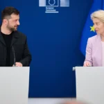 Ο Βολοντίμιρ Ζελένσκι με μαύρο μπουφάν στέκεται σε λευκό βήμα μπροστά σε ουκρανική σημαία και κοιτάζει την Ούρσουλα φον ντερ Λάιεν στο διπλανό βήμα