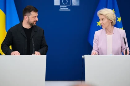 Ο Βολοντίμιρ Ζελένσκι με μαύρο μπουφάν στέκεται σε λευκό βήμα μπροστά σε ουκρανική σημαία και κοιτάζει την Ούρσουλα φον ντερ Λάιεν στο διπλανό βήμα